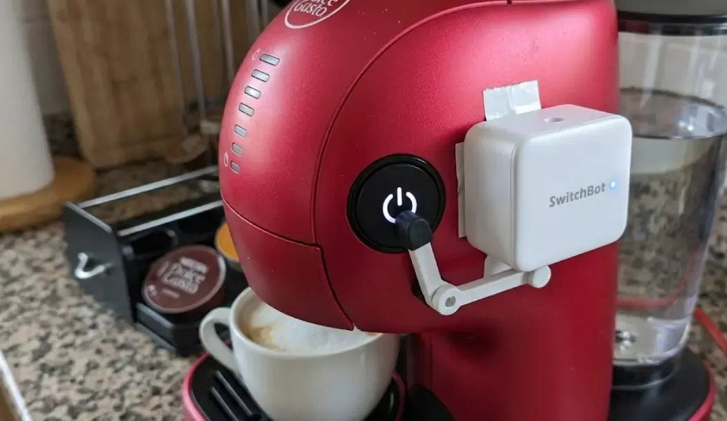 Detalle de un pequeño robot SwitchBot blanco pegado al lado del botón de inicio de una cafetera de cápsulas roja, a punto de presionar el botón de forma automatizada.